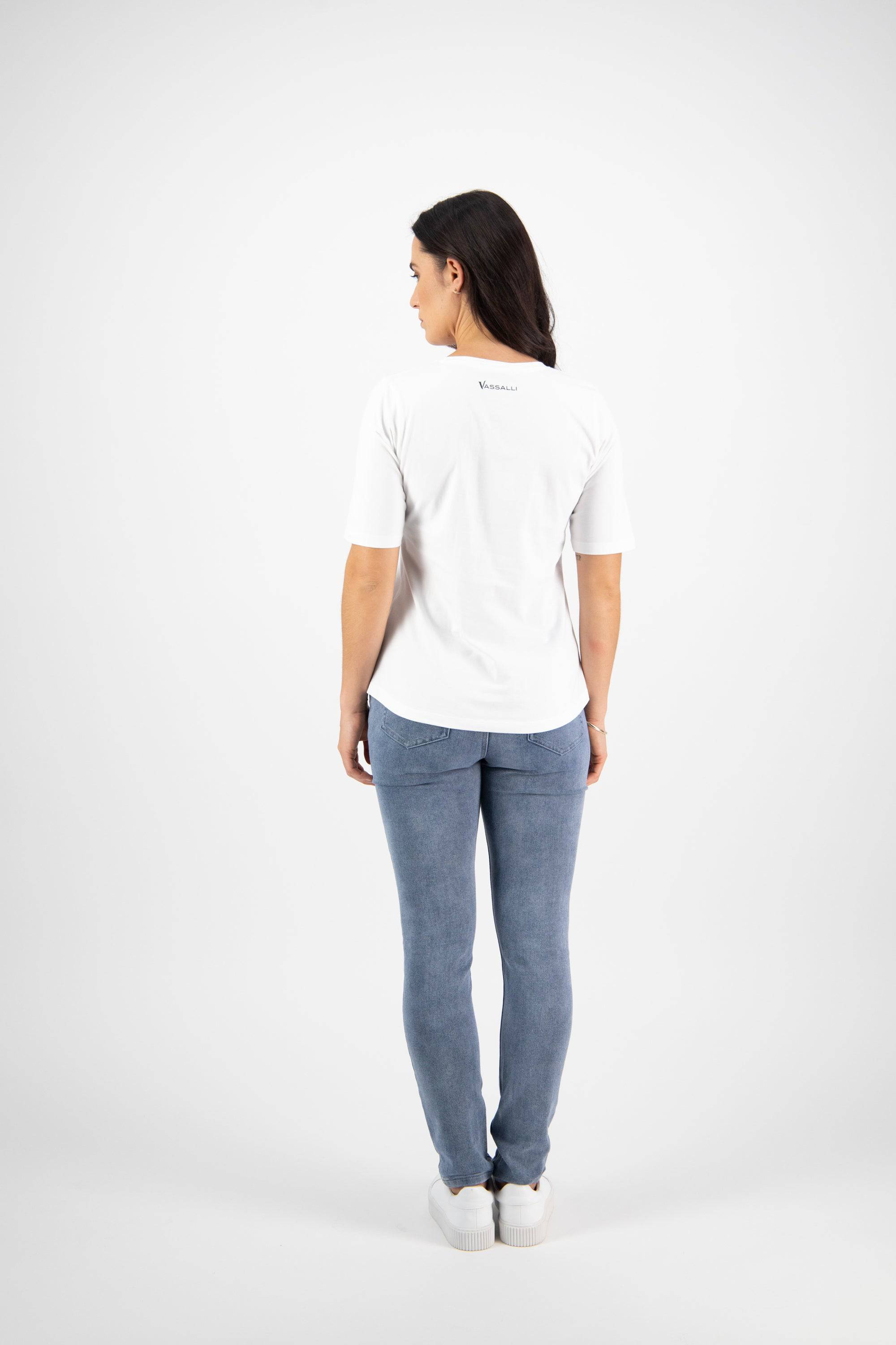 A woman with long dark hair faces away, wearing the Vassalli Base Tee in white and light blue jeans with white sneakers, set against a plain white background.