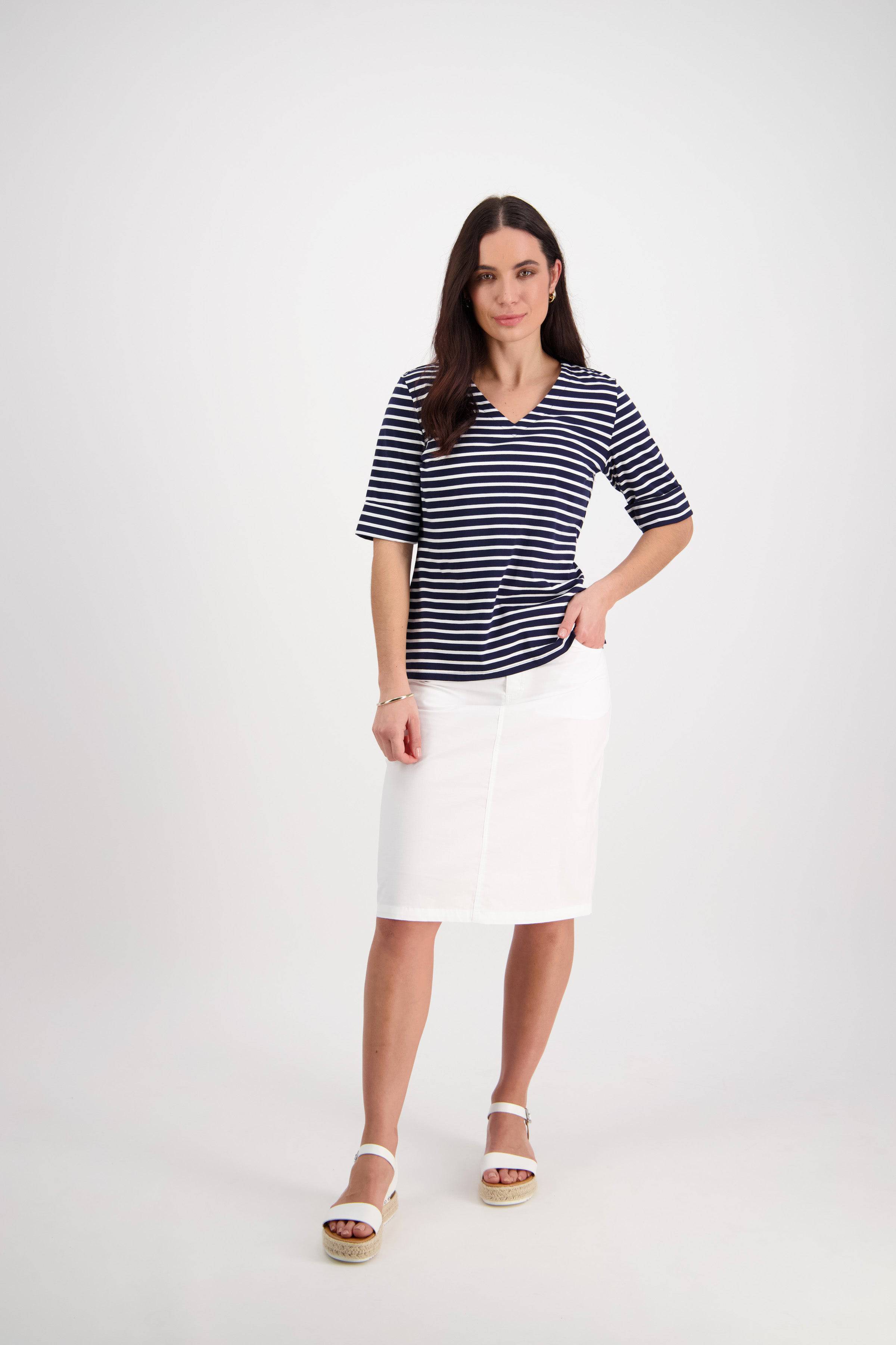 A woman with long brown hair and a neutral expression stands against a white background, wearing a navy and white striped short-sleeve top, Vassalli's Poppy Poplin Skirt in White, and white sandals.
