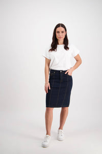 A woman with long dark hair stands facing the camera, wearing a white t-shirt, Vassalli’s Poppy Skirt in Indigo Denim, and white sneakers, one hand in her pocket against a plain white background.