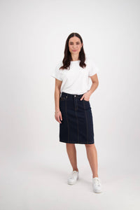 A woman with long dark hair stands against a white background, wearing a white short-sleeve shirt, the Vassalli Poppy Skirt in Indigo Denim with contrast stitching, and white sneakers, one hand tucked into her skirt pocket.