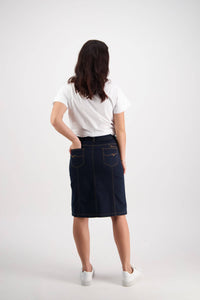 A woman with long brown hair, wearing a white t-shirt, Vassalli’s Poppy Skirt in Indigo Denim, and white sneakers stands facing away against a plain light background.