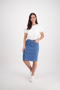 A woman with long dark hair and one hand in her pocket wears a white tee, white sneakers, and the Vassalli Poppy Skirt in light denim against a plain white background.