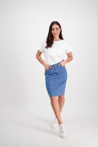 A woman with long dark hair stands against a white background, wearing Vassalli’s Poppy Skirt in Light Denim, a white T-shirt, and white sneakers. She rests her hands in her skirt pockets and smiles slightly.