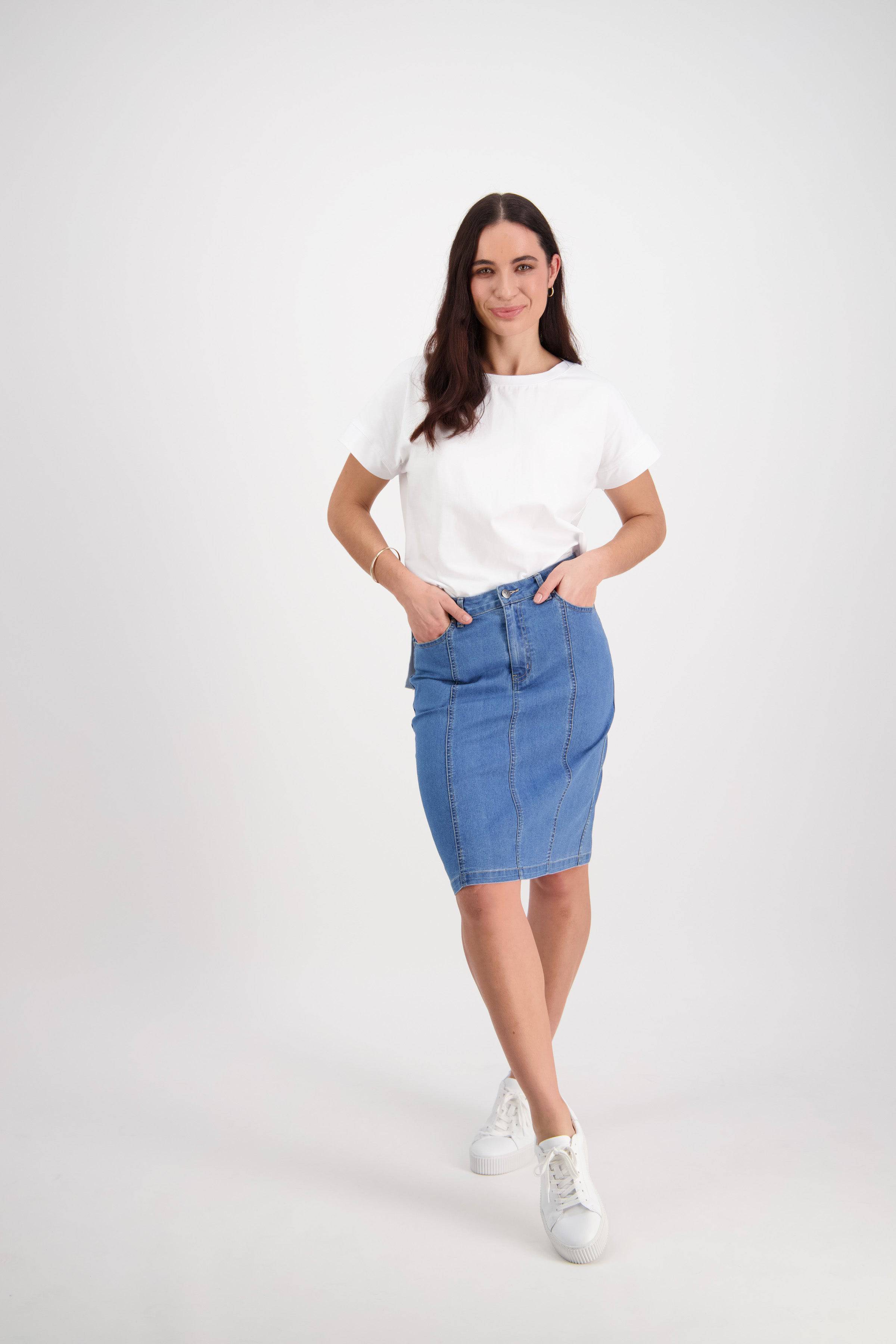 A woman with long dark hair stands against a white background, wearing Vassalli’s Poppy Skirt in Light Denim, a white T-shirt, and white sneakers. She rests her hands in her skirt pockets and smiles slightly.