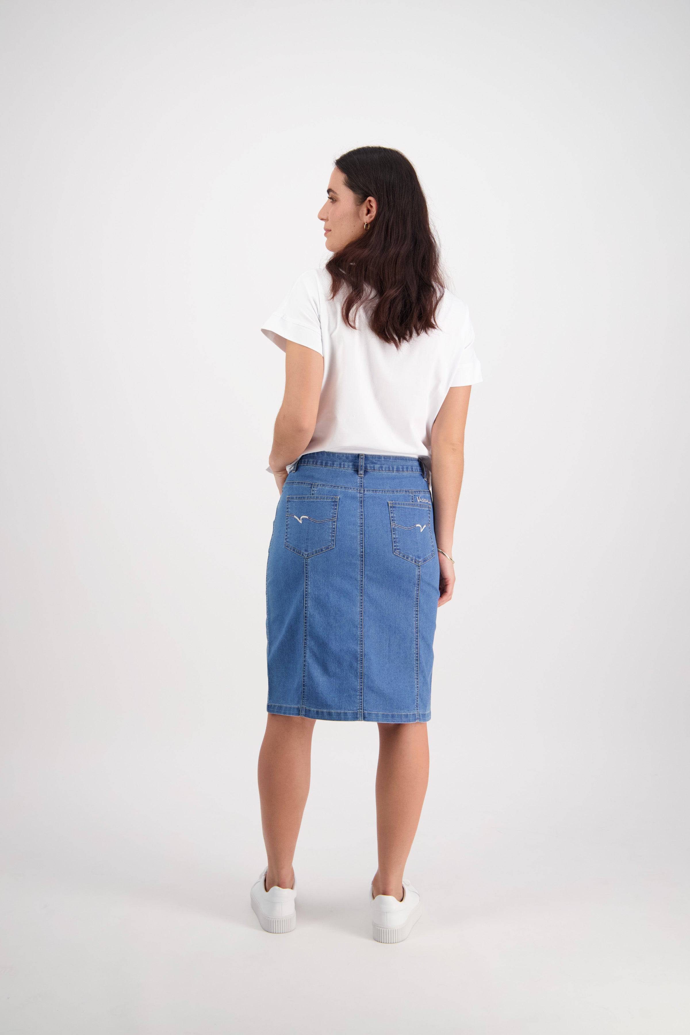 A woman with dark hair faces away against a white background, wearing a white tee, white sneakers, and the Vassalli Poppy Skirt in light denim with contrast stitch detailing.