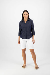A woman smiles against a plain white background, wearing the Vassalli Isla Shirt in Ink— a navy slim fit, long sleeve shirt—paired with white knee-length shorts and beige sandals.