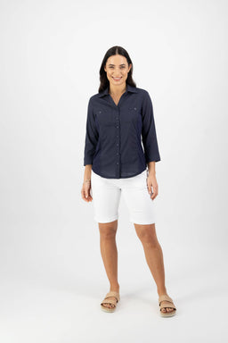 A woman smiles against a plain white background, wearing the Vassalli Isla Shirt in Ink— a navy slim fit, long sleeve shirt—paired with white knee-length shorts and beige sandals.