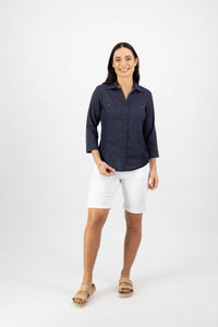 A woman with long dark hair smiles against a white background, wearing the Vassalli Isla Shirt in Ink—a navy 100% cotton collared button-up with 3/4 sleeves—paired with white knee-length shorts and beige sandals in a relaxed pose.