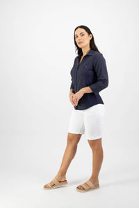 A woman with long dark hair stands against a white background wearing the Vassalli Isla Shirt - Ink, paired with white knee-length shorts and tan sandals. She poses relaxedly, gazing slightly past the camera.