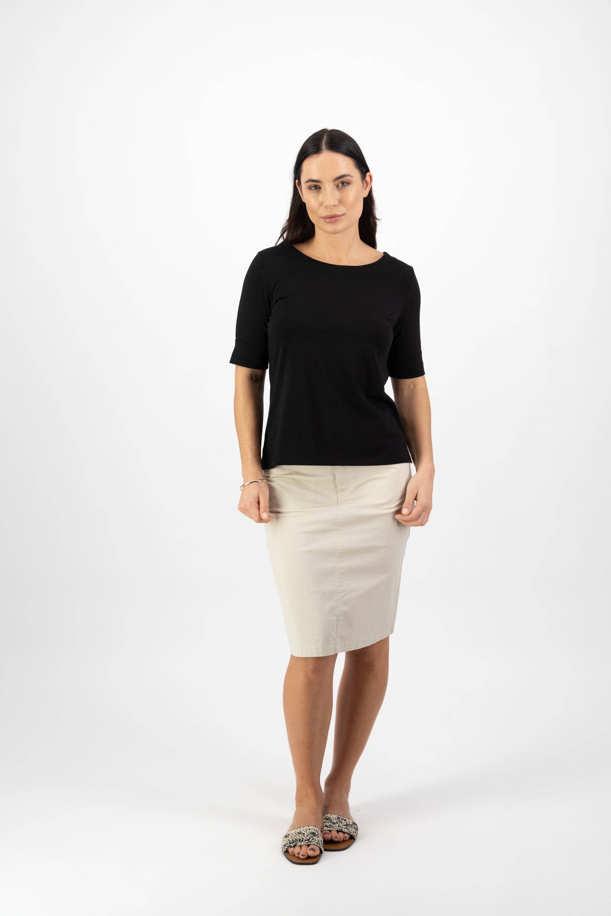 A woman with long dark hair stands against a white background, wearing the Vassalli Boat Neck Top in black and a beige skirt. She faces forward with a neutral expression, arms relaxed at her sides.