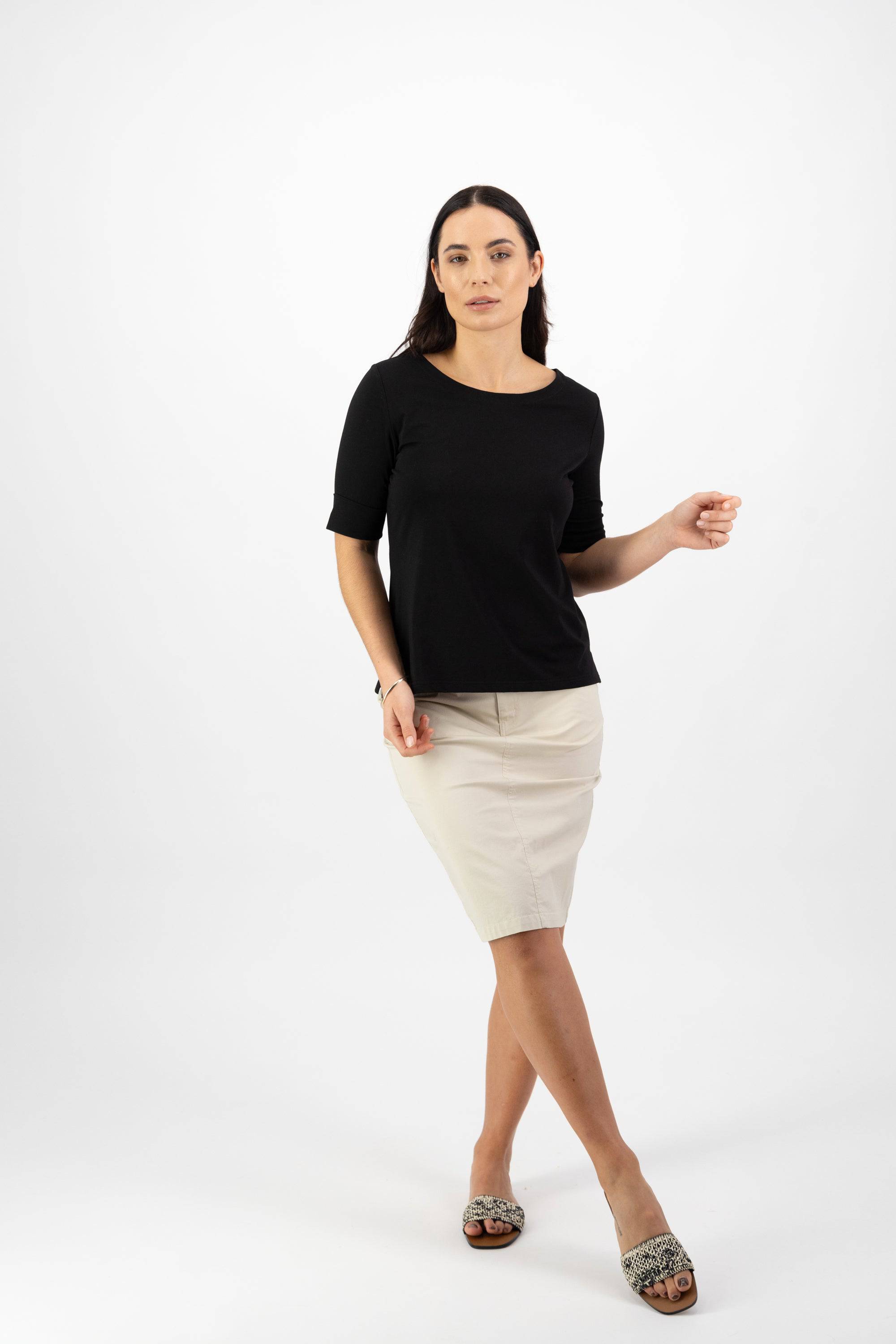A woman with long dark hair and a neutral expression wears the Vassalli Boat Neck Top in black against a white background. She pairs it with a light beige knee-length skirt and slip-on shoes, one arm slightly raised.
