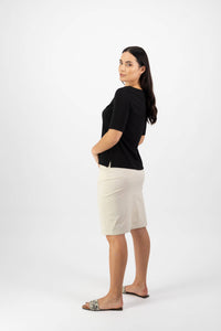 A woman with long dark hair poses sideways against a white background, wearing the Vassalli Boat Neck Top in black, paired with a cream knee-length skirt and slip-on sandals.