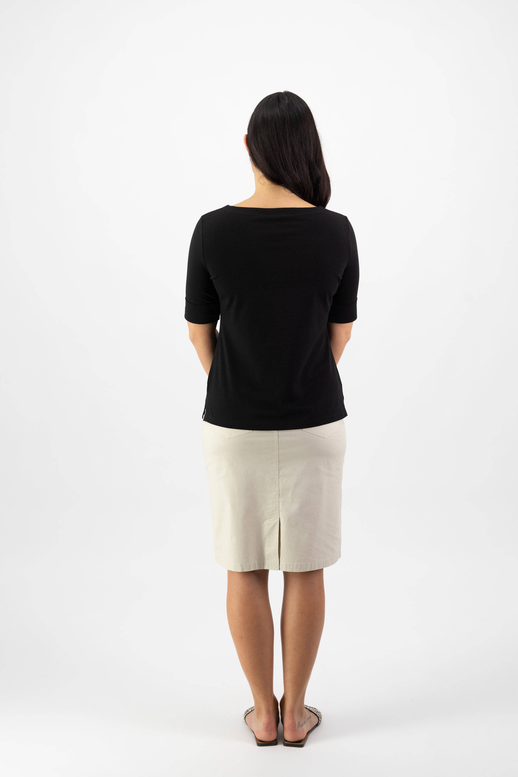 A woman with long dark hair models the Vassalli Boat Neck Top in black, paired with a beige knee-length skirt and sandals, as she stands facing away from the camera against a plain white background.