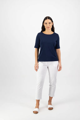 A woman with long dark hair stands against a white background, wearing the Vassalli Boat Neck Top - Navy in a relaxed fit, paired with white cropped pants and sandals. She faces forward with a neutral expression.