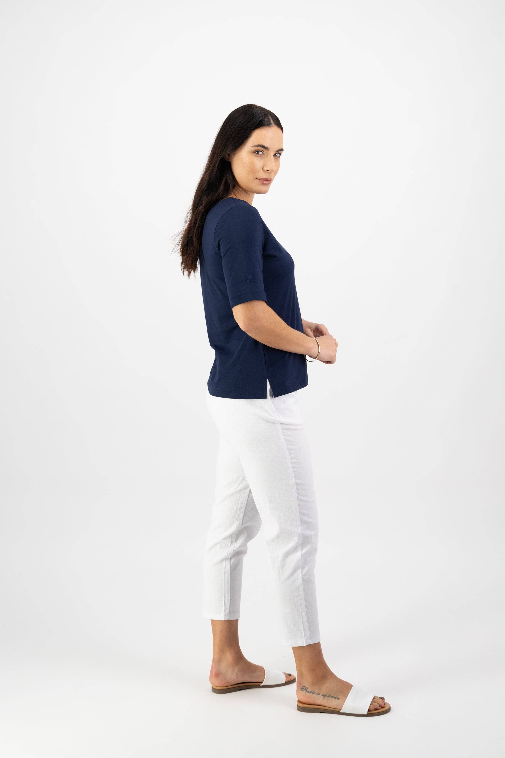 A woman with long dark hair stands sideways, wearing the Vassalli Boat Neck Top - Navy in a relaxed fit, paired with white cropped pants and white slide sandals against a plain white background. She looks at the camera with a neutral expression.