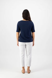 A woman faces away from the camera wearing the Vassalli Boat Neck Top - Navy, a relaxed fit cotton-elastane jersey, styled with white pants and sandals. Her dark hair is down against a plain white background.