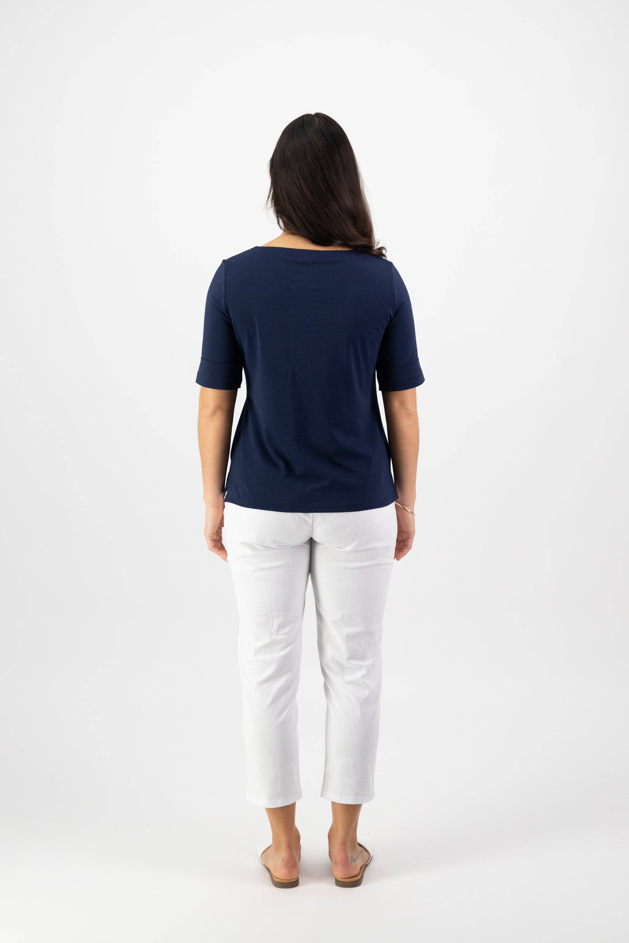 A woman faces away from the camera wearing the Vassalli Boat Neck Top - Navy, a relaxed fit cotton-elastane jersey, styled with white pants and sandals. Her dark hair is down against a plain white background.