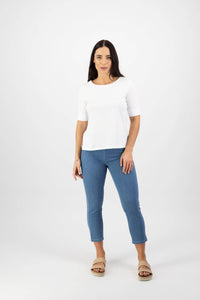 A woman with long dark hair and a neutral expression stands facing forward against a white background, wearing Vassalli's Boat Neck Top in white paired with blue jeans and beige sandals.