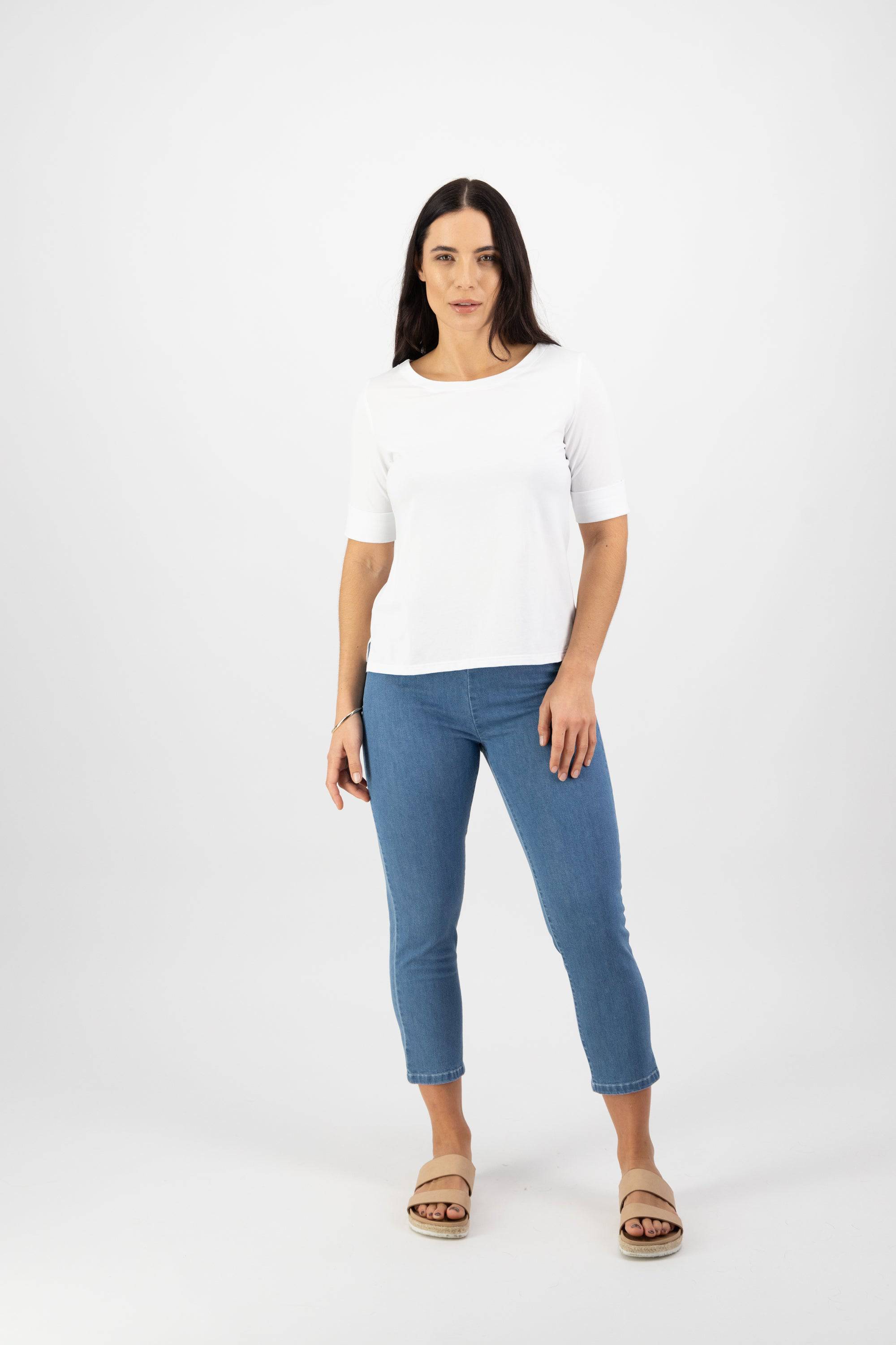 A woman with long dark hair and a neutral expression stands facing forward against a white background, wearing Vassalli's Boat Neck Top in white paired with blue jeans and beige sandals.