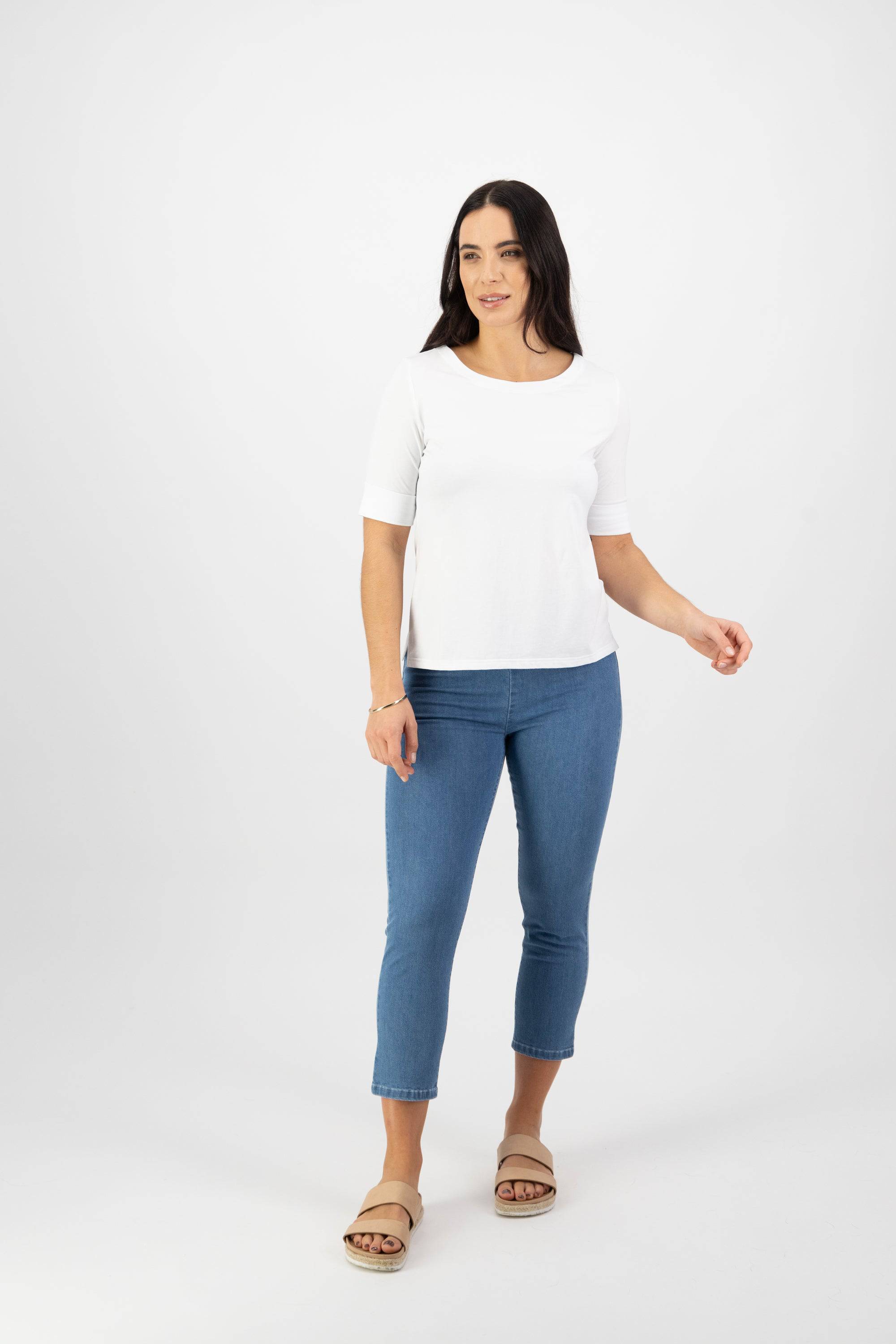 A woman with long dark hair wears the Vassalli Boat Neck Top in white, paired with blue cropped jeans and beige sandals. She stands against a plain white background, looking slightly left with a relaxed expression.