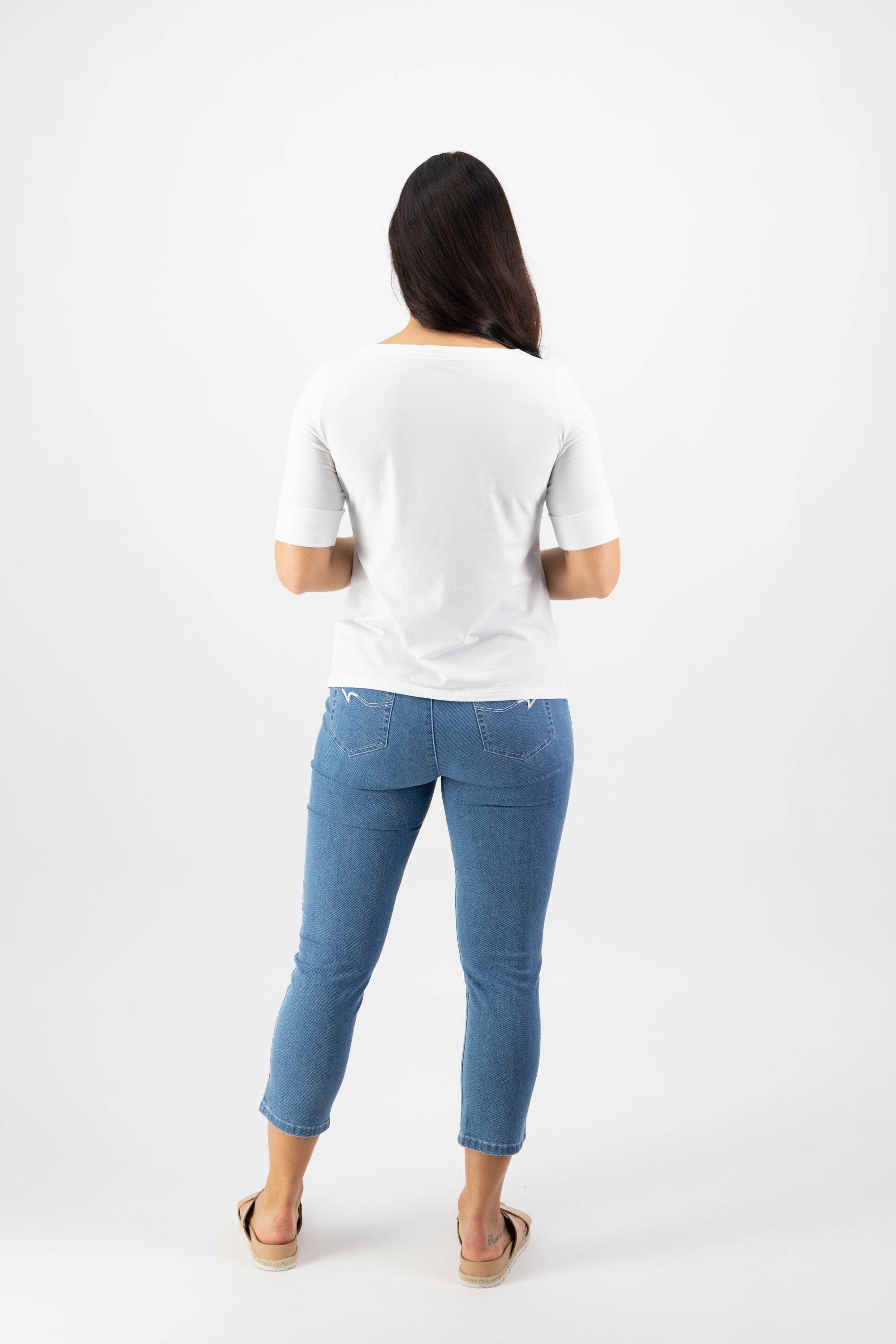 A woman faces away from the camera wearing the Vassalli Boat Neck Top in white, paired with blue skinny jeans and beige slip-on shoes, all set against a plain white background.