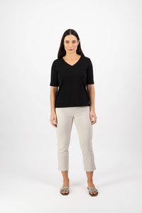 A woman with long dark hair stands facing forward wearing the Vassalli V-Neck Top in black, paired with light beige cropped pants and patterned slip-on shoes, against a plain white background.