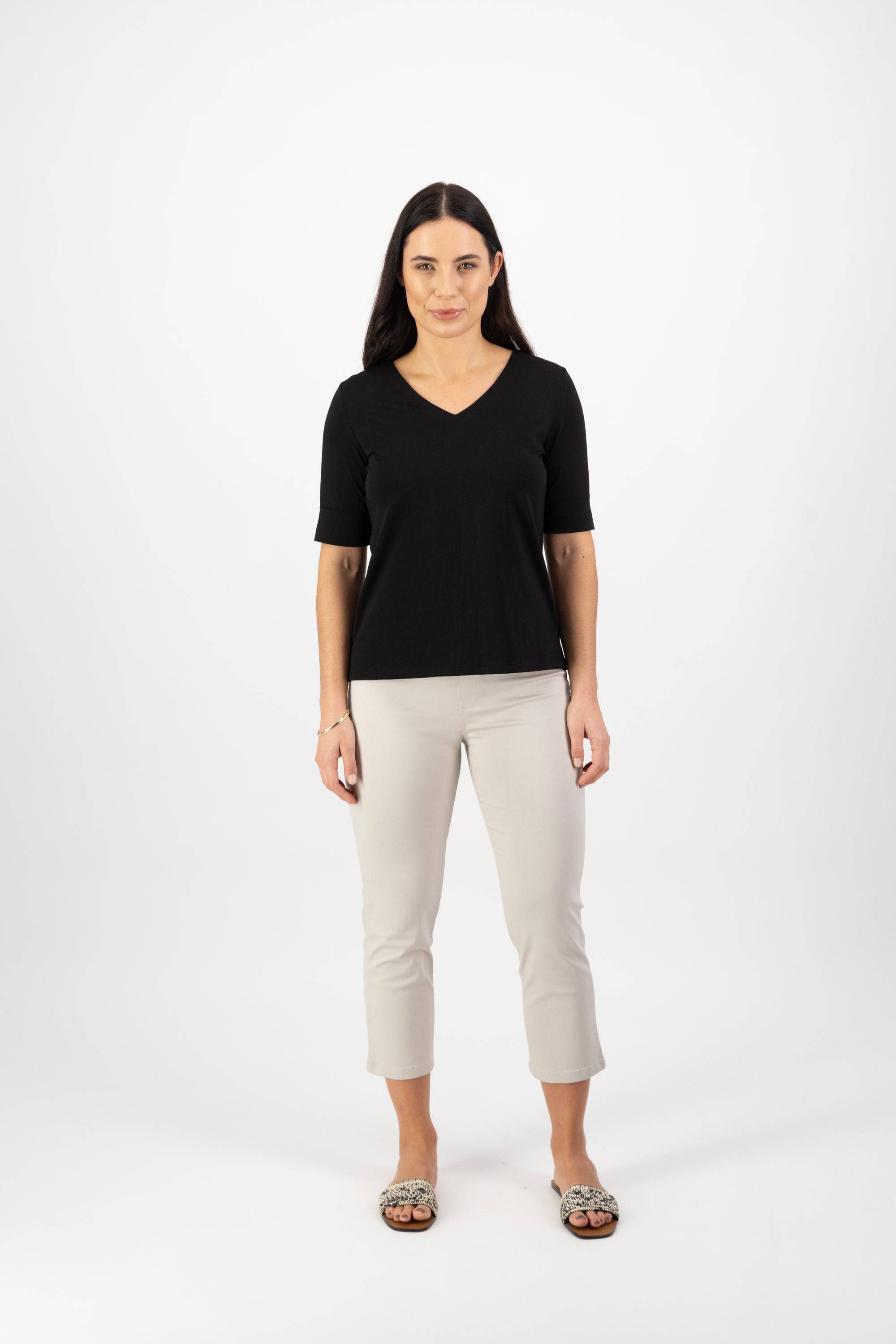 A woman with long dark hair stands facing forward wearing the Vassalli V-Neck Top in black, paired with light beige cropped pants and patterned slip-on shoes, against a plain white background.