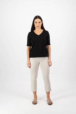 A woman with long dark hair stands facing forward wearing the Vassalli V-Neck Top in black, paired with light beige cropped pants and patterned slip-on shoes, against a plain white background.