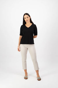 A woman with long dark hair stands against a white background, wearing the Vassalli V-Neck Top in black. She pairs it with light beige cropped pants and open-toed sandals, posing with one arm behind her back and a neutral expression.