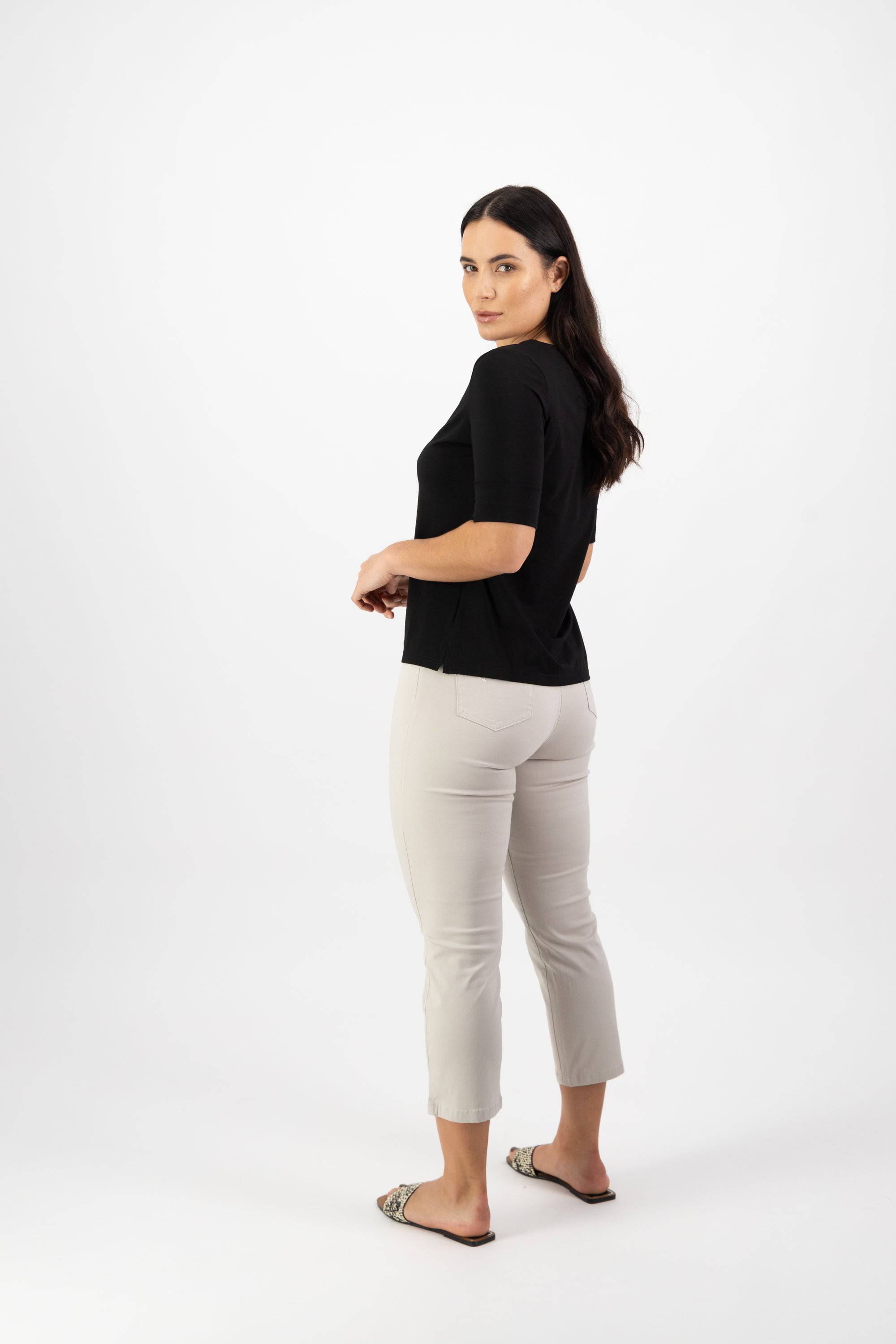 A woman with long dark hair stands against a white background, looking over her shoulder. She wears the Vassalli V-Neck Top in black, paired with light pants and sandals.