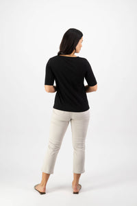 A woman faces away from the camera, wearing the Vassalli V-Neck Top - Black in stretch cotton jersey, paired with light beige relaxed-fit cropped pants and sandals, set against a plain white background.