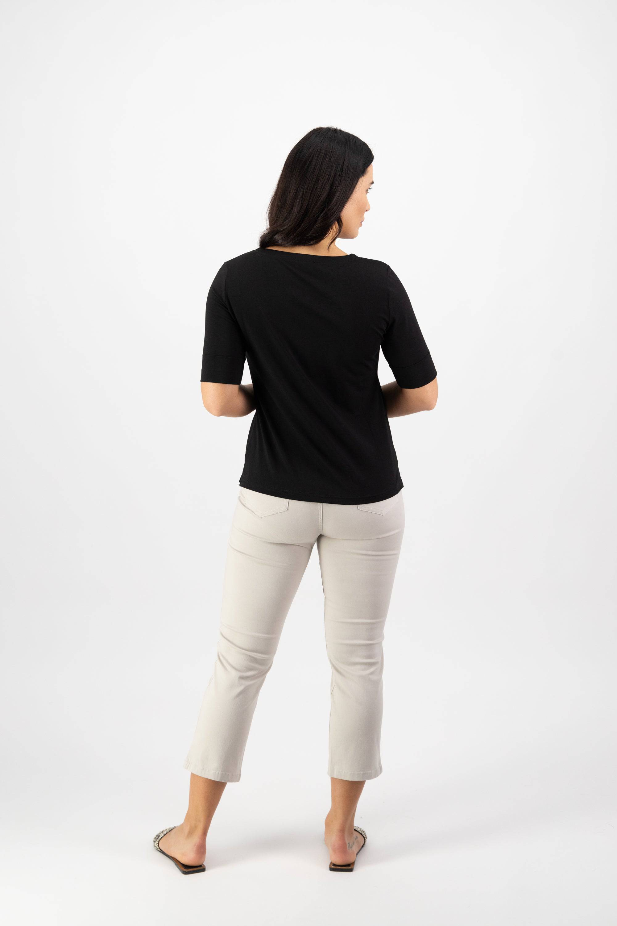 A woman faces away from the camera, wearing the Vassalli V-Neck Top - Black in stretch cotton jersey, paired with light beige relaxed-fit cropped pants and sandals, set against a plain white background.