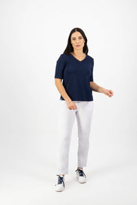 A woman with long dark hair stands against a white background wearing the Vassalli V-Neck Top - Navy in stretch cotton jersey, paired with white pants and black-and-white sneakers. She has a neutral expression and one hand slightly raised.