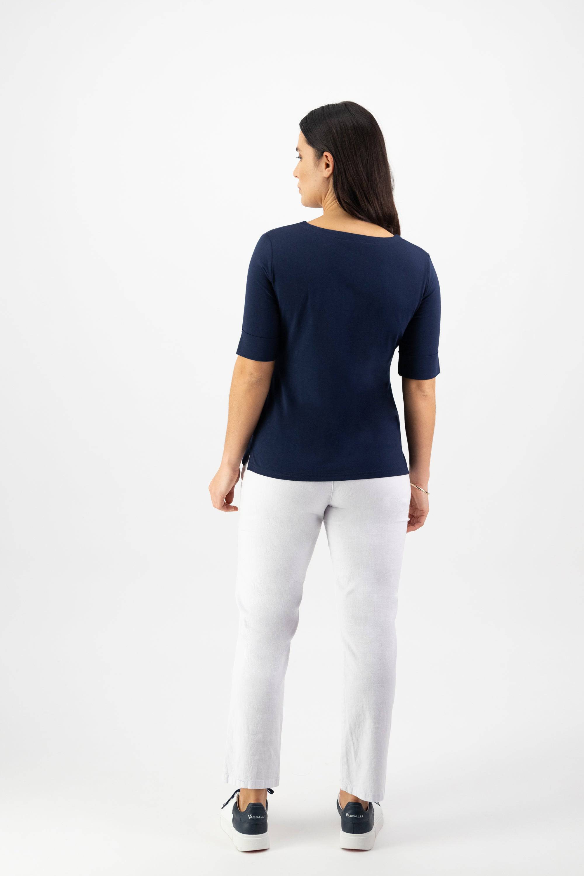A woman with long dark hair faces away, wearing the Vassalli V-Neck Top - Navy in stretch cotton jersey, paired with white pants and dark sneakers, set against a plain white background.