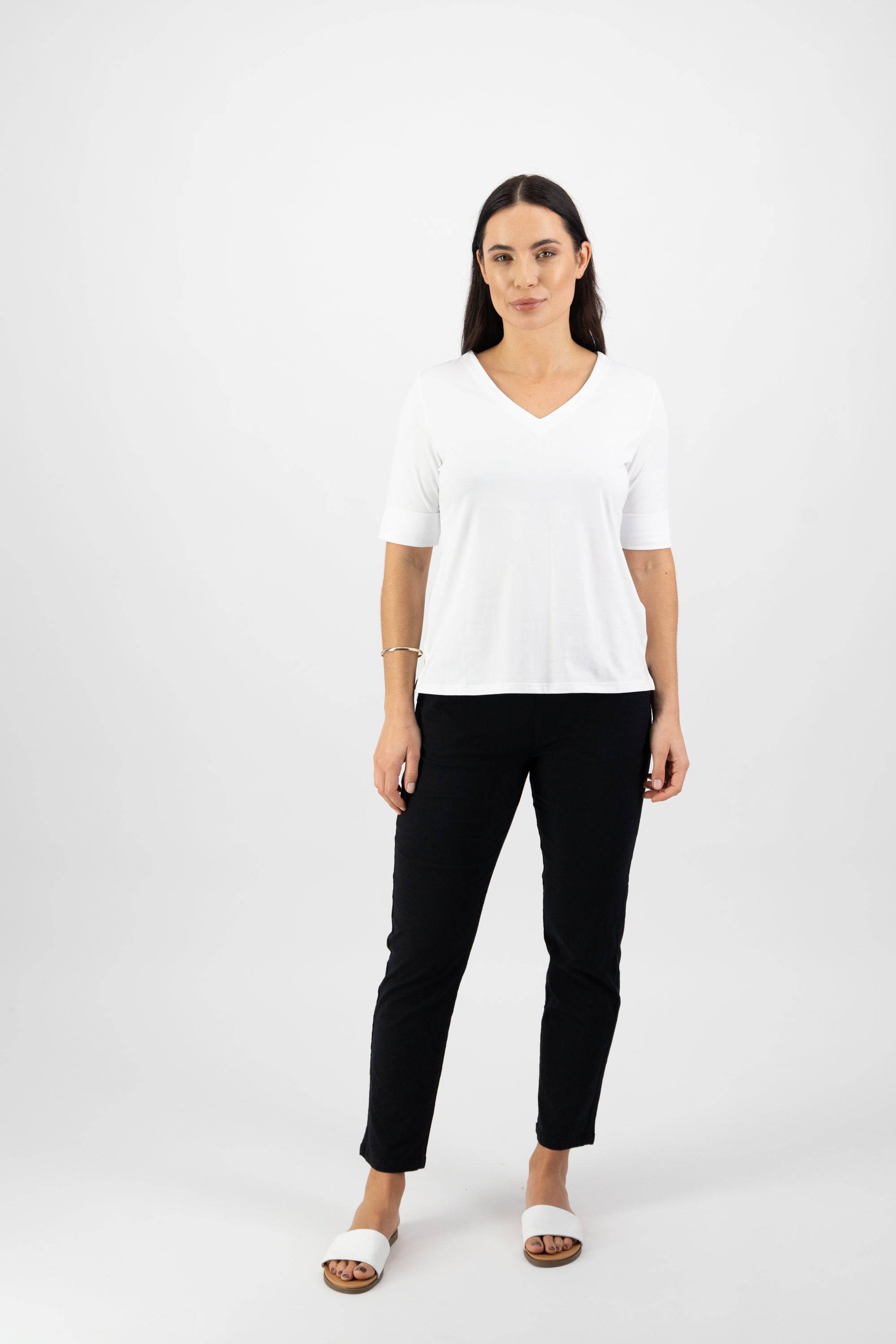 A woman with long dark hair stands against a white background, wearing the Vassalli V-Neck Top in white and black pants. She pairs her outfit with white slide sandals and a bracelet on her right wrist, looking straight at the camera neutrally.