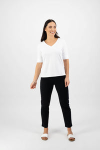 A woman with long dark hair smiles against a plain white background, wearing the Vassalli V-Neck Top in white along with black pants and white sandals.