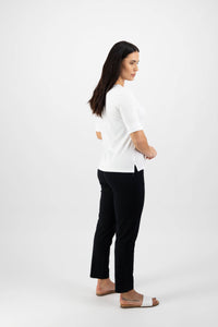 A woman with long dark hair poses sideways against a white background, wearing the Vassalli V-Neck Top in white, paired with black pants and beige sandals. Her arms are loosely crossed.