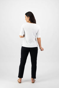 A person with long dark hair faces away from the camera, wearing the Vassalli V-Neck Top in white, black pants, and beige flat shoes against a plain white background.