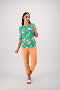 A woman with long dark hair poses against a plain background, wearing the Carmen Top - Lush by Vassalli—a green floral 3/4 sleeve knit with a relaxed fit—paired with light orange pants and white sandals, looking to the side with one hand raised.
