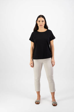 A woman with long dark hair stands facing forward, wearing the Vassalli Thea Top in black, light beige cropped pants, and leopard-print slip-on shoes against a plain white background.
