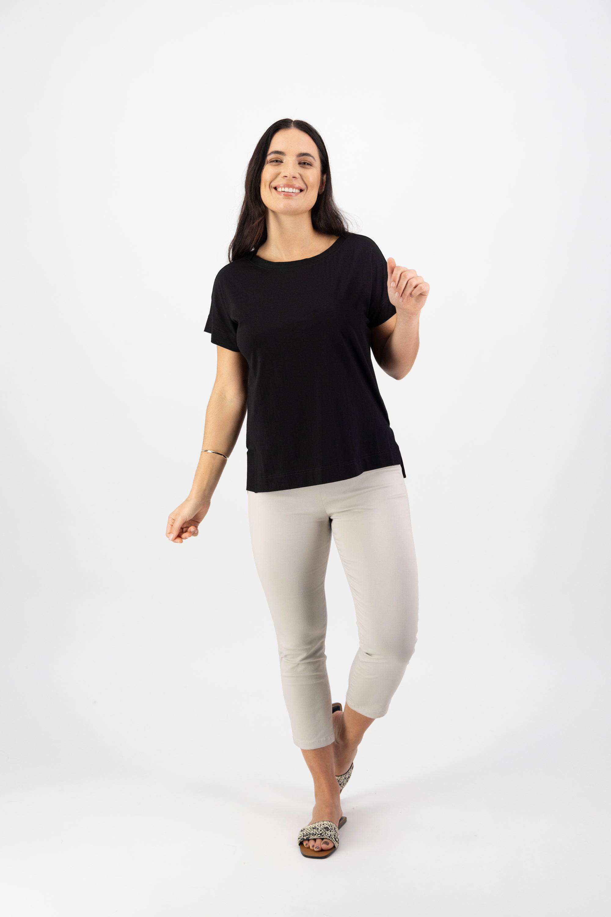 A woman with long dark hair smiles and poses against a white background, wearing the Vassalli Thea Top in black paired with light cropped pants and sandals.