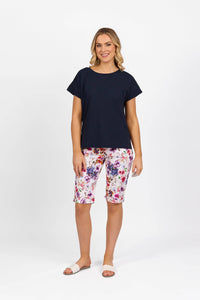 A woman stands against a white background, smiling in the Vassalli Thea Top - Ink, colorful floral knee-length shorts, and white slide sandals. Her arms rest relaxed by her sides.
