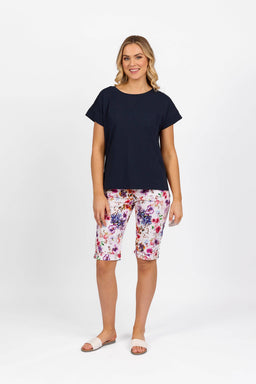 A woman stands against a white background, smiling in the Vassalli Thea Top - Ink, colorful floral knee-length shorts, and white slide sandals. Her arms rest relaxed by her sides.