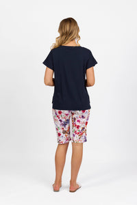 A woman stands with her back to the camera, wearing the Vassalli Thea Top in Ink, floral-patterned knee-length shorts, and flip-flops against a plain white background.