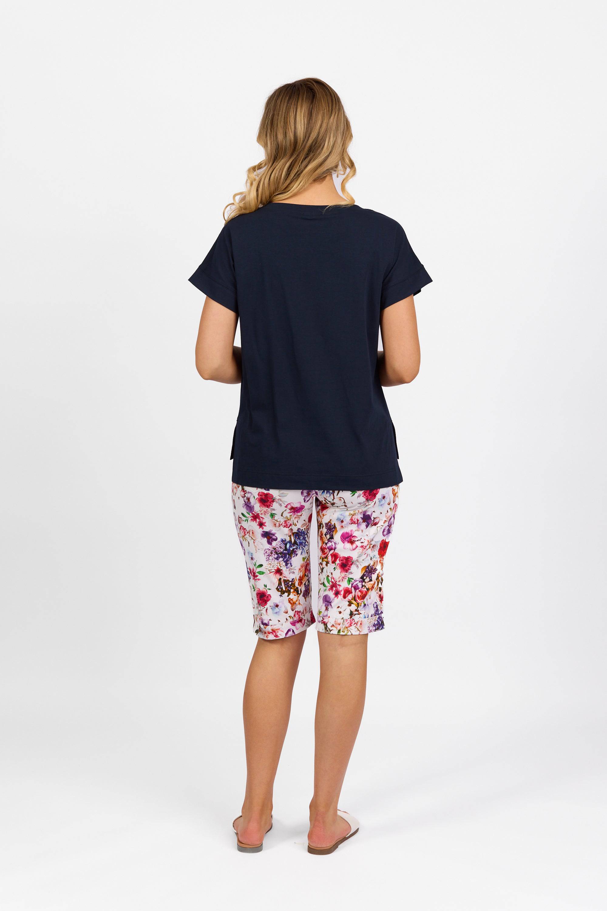 A woman stands with her back to the camera, wearing the Vassalli Thea Top in Ink, floral-patterned knee-length shorts, and flip-flops against a plain white background.