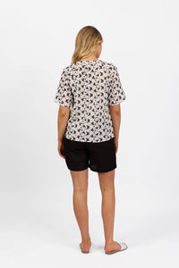 A woman faces away from the camera, wearing the Vassalli V-Neck Top - Floral, paired with black shorts and flat sandals, set against a plain white background.