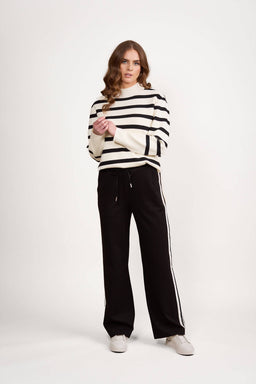 A woman in a relaxed pose stands against a plain white background, wearing Vassalli’s Weekend Pant - Black 2.0 with a side stripe panel, a white sweater with black stripes, and white sneakers. Her hair is wavy.
