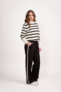 A woman with long wavy hair and a neutral expression poses against a white background, wearing Vassalli's Weekend Pant - Black 2.0 with a white side stripe, a black and white striped sweater, and white sneakers.