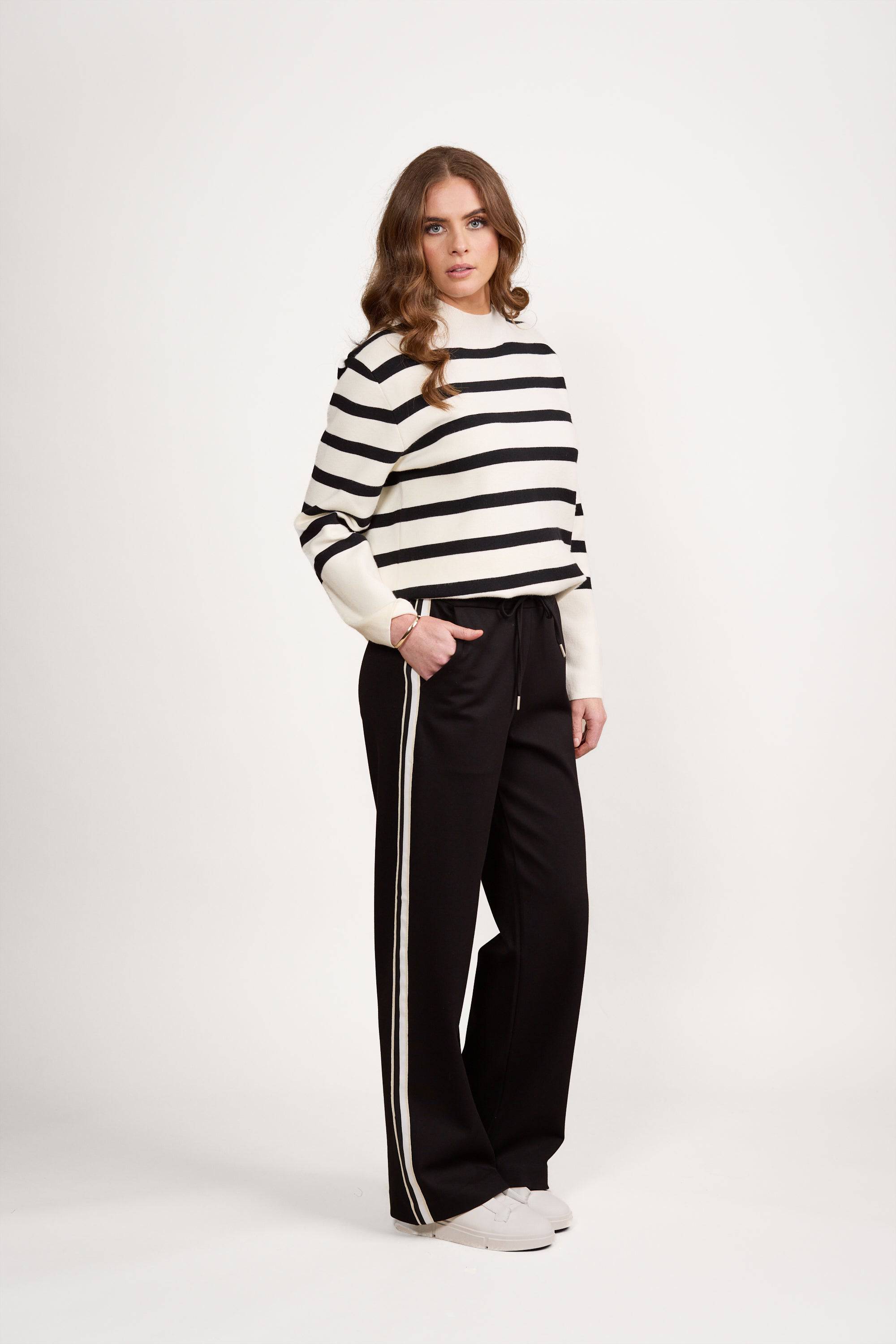 A woman with long wavy hair and a neutral expression poses against a white background, wearing Vassalli's Weekend Pant - Black 2.0 with a white side stripe, a black and white striped sweater, and white sneakers.