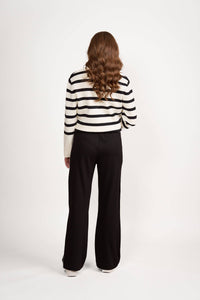 A person with long, wavy brown hair, facing away, wears a white sweater with black stripes and Vassalli’s Weekend Pant - Black 2.0, loose wide-leg stretch pants, against a plain white background.